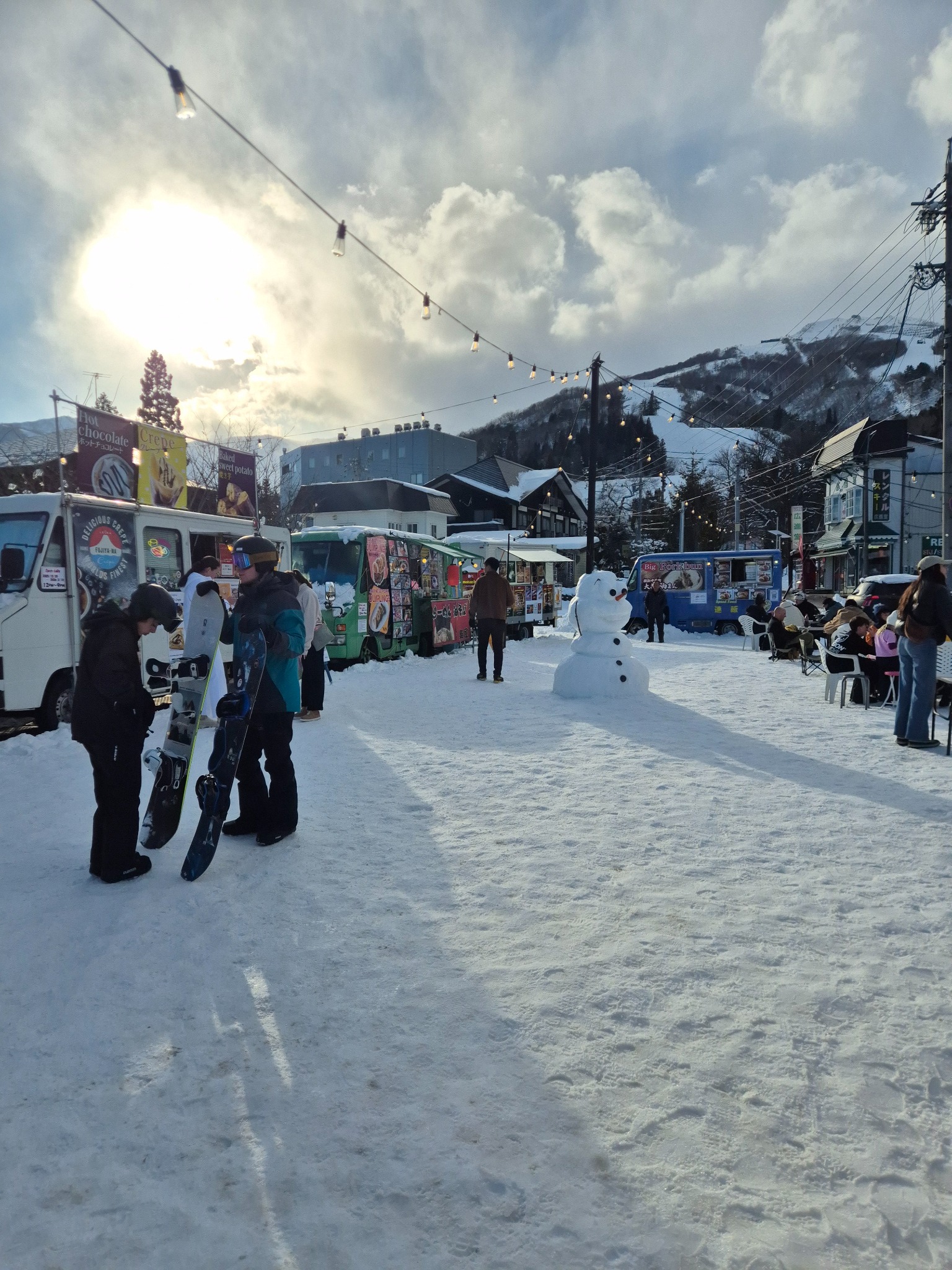 Skiers beside food stalls and a snowman in a snowy Japanese mountain village